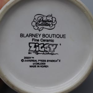 May include: A white ceramic mug with a black handle and the text "But First, Coffee" in a bold, black font. The mug is filled with a dark liquid, likely coffee, and sits on a light-colored surface.