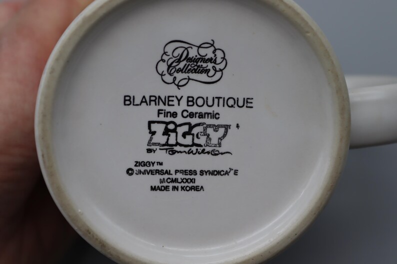 May include: A white ceramic mug with a black handle and the text "But First, Coffee" in a bold, black font. The mug is filled with a dark liquid, likely coffee, and sits on a light-colored surface.