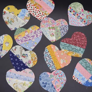 May include: A collection of fabric heart shapes in various sizes and patterns. The hearts are made from patchwork fabric with floral, polka dot, and geometric designs in shades of pink, blue, yellow, and green. The hearts are arranged on a dark gray surface.