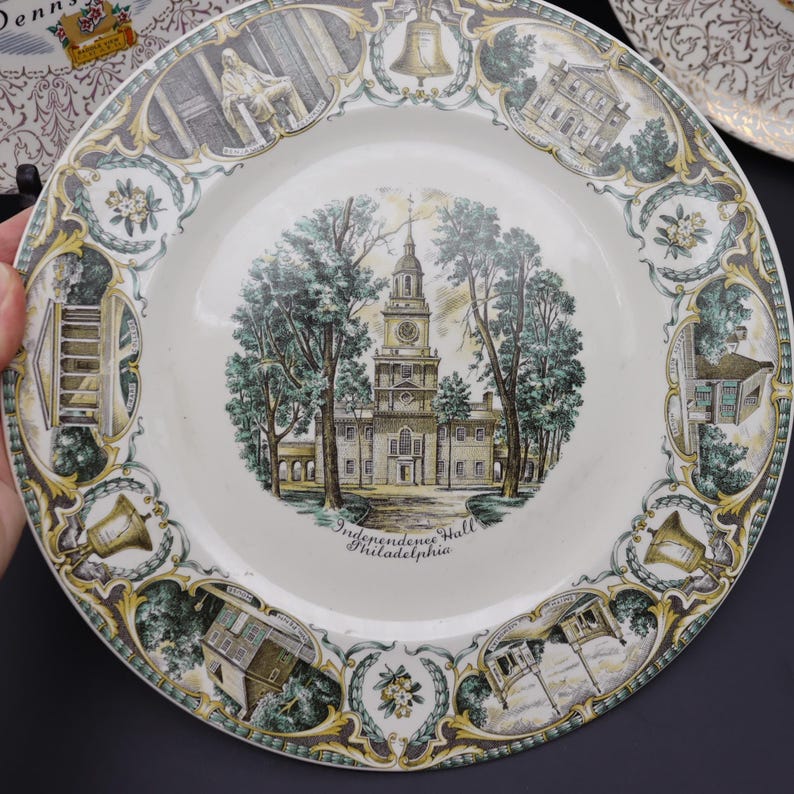 K&ouml;nnte beinhalten: Ein dekorativer Vintage-Teller mit der Independence Hall in Philadelphia. Der Teller hat eine cremefarbene Mitte mit einer detaillierten Illustration des Geb&auml;udes, umgeben von einem dekorativen Rand mit historischen Motiven in Gr&uuml;n, Gold und Grau.