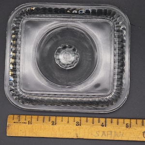 Puede incluir: Tapa de vidrio transparente de forma cuadrada con un borde decorativo y estriado. La tapa tiene una hendidura central circular y un pequeño botón elevado en el centro. Una regla de madera con medidas en pulgadas está en primer plano. Dimensiones: aprox. 10 cm.