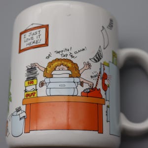 May include: White ceramic mug with a cartoon illustration of a busy office worker. The mug features the text "I JUST LOVE IT HERE!" and other sound effects. The design includes a desk, phone, and stacks of paperwork.