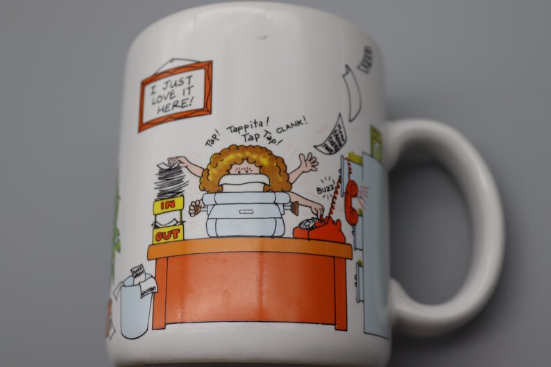 May include: White ceramic mug with a cartoon illustration of a busy office worker. The mug features the text "I JUST LOVE IT HERE!" and other sound effects. The design includes a desk, phone, and stacks of paperwork.