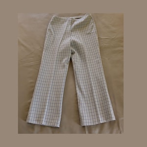 May include: A pair of vintage, high-waisted pants with a white and blue plaid pattern. The pants have a flared leg design and are made of a lightweight fabric. The pants are displayed on a neutral background.