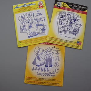 May include: Three Aunt Martha's hot iron transfer packets featuring blue line art designs. The top left packet is titled Clancy the Clown, the top right is Nursery Rhyme Crib Spread, and the bottom packet displays Sunbonnet Sue and Overall Bill.