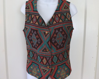 May include: A colorful, patterned vest with a V-neck and wooden buttons. The vest features a geometric design in teal, red, brown, and gold. The vest is displayed on a white mannequin.