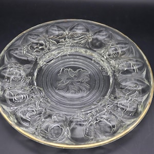May include: A round clear glass serving plate featuring a central embossed floral motif surrounded by a ring of twelve individual egg-shaped indentations. The outer edge of the plate is finished with a thin gold-colored metallic trim.
