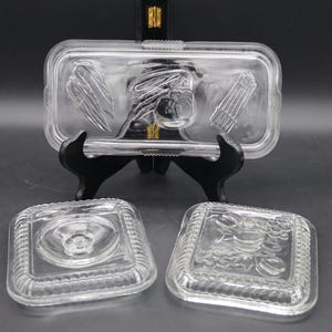 May include: Clear glass serving dish with a rectangular lid featuring embossed designs of a bird, feathers, and asparagus. Two square glass dishes are also visible, one with a floral design and the other with a circular design.