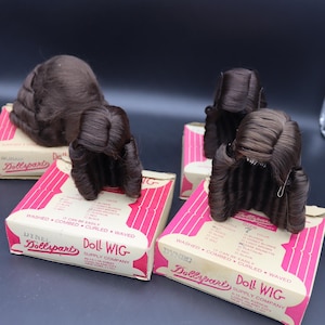 Op de afbeelding: Verschillende bruine poppenpruiken worden getoond op hun originele dozen. De dozen zijn wit met roze strepen en de tekst "Doll Wig" en "Dynel Dollspart Supply Company". De pruiken hebben een krullende stijl.