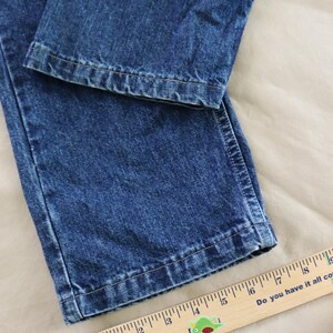 Op de afbeelding: Close-up van een blauwe spijkerbroek. De jeans is opgevouwen en toont de zoom en de textuur van de denim. Een houten liniaal met centimeter markeringen is zichtbaar, met de tekst "Do you have it all covered?"