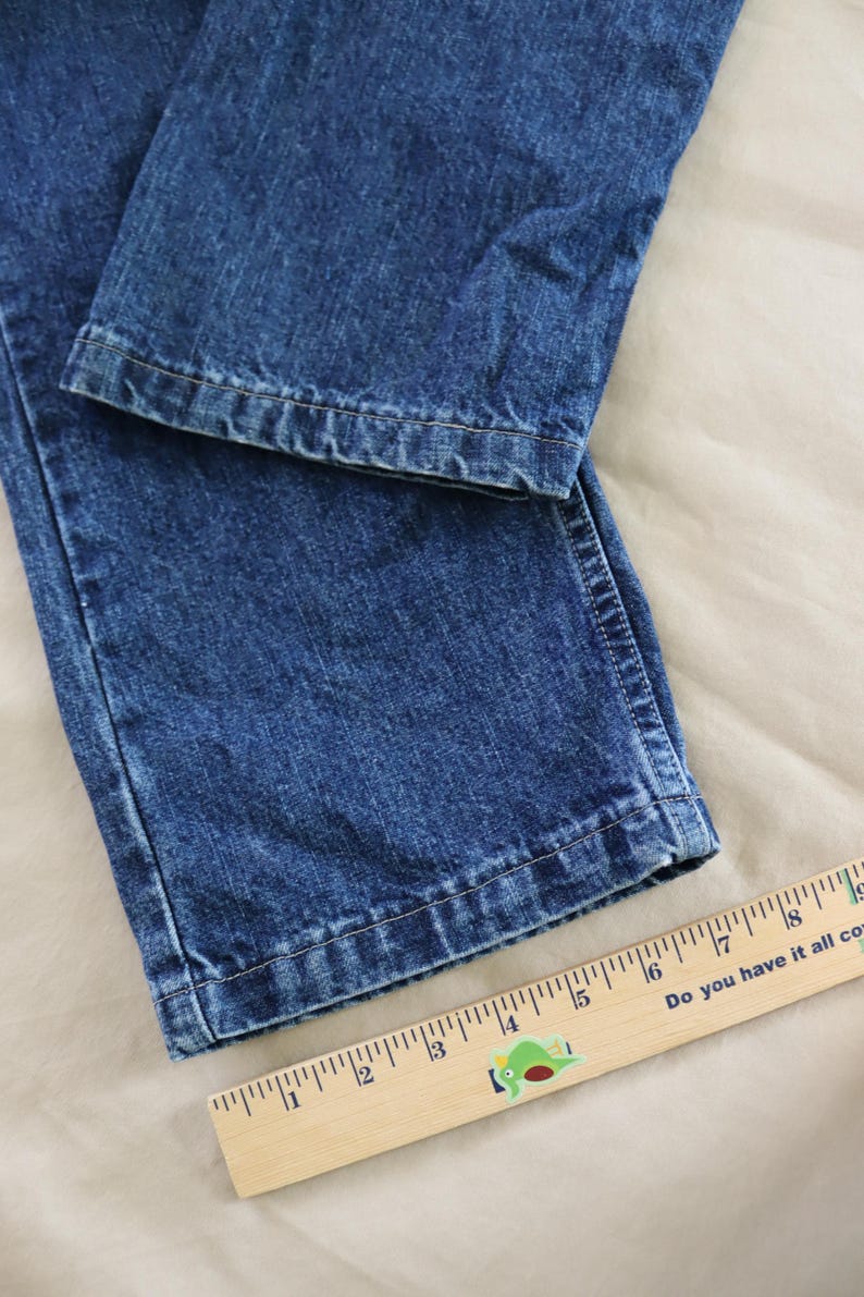 Op de afbeelding: Close-up van een blauwe spijkerbroek. De jeans is opgevouwen en toont de zoom en de textuur van de denim. Een houten liniaal met centimeter markeringen is zichtbaar, met de tekst "Do you have it all covered?"