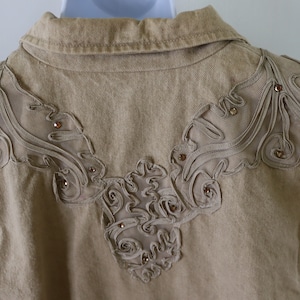 Puede incluir: Una chaqueta vaquera de color marrón claro con un diseño decorativo en la espalda. El diseño está hecho de una tela más clara y presenta un patrón floral con pedrería.