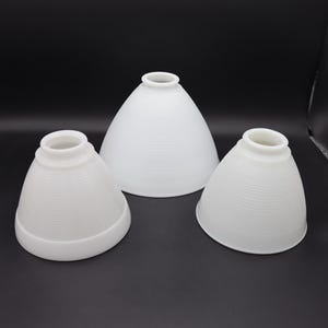 以下が含まれることがあります： テクスチャ加工された水平リブデザインの、白い円錐形のランプシェードが3つ。シェードは丸みを帯びたベースと上部に小さな開口部があります。シェードはさまざまなサイズで、暗い表面に配置されています。