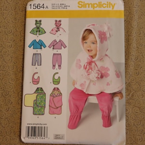 May include: Simplicity sewing pattern 1564A featuring baby clothing designs. The pattern includes options for a hooded cape, wrap top, pants, bib, and a baby bunting. The image shows a baby wearing a pink hooded cape and pants.