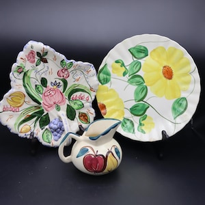 May include: A collection of hand-painted ceramic tableware. Includes a leaf-shaped dish with pink roses and green leaves, a round plate with yellow flowers, and a small pitcher decorated with fruit.