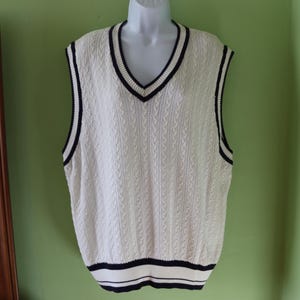 May include: Cream cable knit sweater vest with a V-neck and navy trim. The sleeveless vest is displayed on a white mannequin. The bottom hem and edges of the vest are trimmed with navy blue stripes.