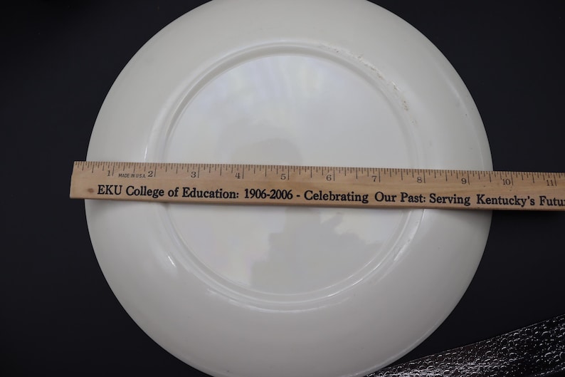 K&ouml;nnte beinhalten: Ein wei&szlig;er, runder Teller mit gl&auml;nzendem Finish. Ein Holzlineal liegt auf dem Teller und misst etwa 25 cm im Durchmesser. Der Text "EKU College of Education" ist auf dem Lineal zu sehen.