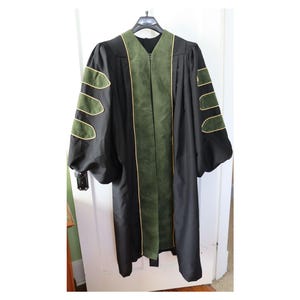 Puede incluir: Una toga académica negra con detalles verdes y ribetes dorados. La toga tiene un panel central verde y paneles verdes en las mangas, todos con ribetes dorados. Colgada de una percha negra.