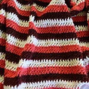 May include: A handmade crochet blanket with a striped pattern. The blanket features alternating stripes of white, coral, and dark brown. The texture of the crochet stitches is visible, creating a cozy and textured appearance. This blanket is suitable for home decor.