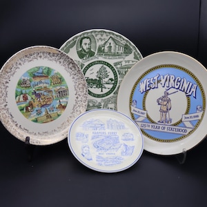 Puede incluir: Una colección de platos decorativos con temas de Virginia Occidental. Los platos muestran varios diseños, incluyendo mapas, figuras históricas y símbolos estatales. La paleta de colores incluye dorado, azul, verde y blanco, con detalles intrincados y texto.