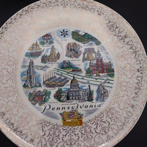 K&ouml;nnte beinhalten: Ein dekorativer Teller mit einer Karte der Sehensw&uuml;rdigkeiten von Pennsylvania. Der Teller hat einen wei&szlig;en und goldenen Filigranrand und ein zentrales Bild mit Illustrationen von Geb&auml;uden und dem Wort "Pennsylvania".