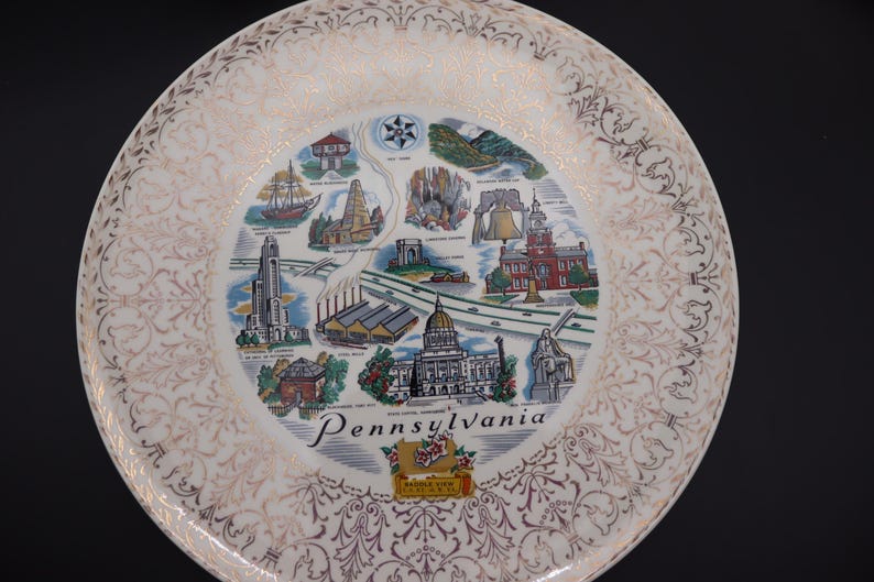 K&ouml;nnte beinhalten: Ein dekorativer Teller mit einer Karte der Sehensw&uuml;rdigkeiten von Pennsylvania. Der Teller hat einen wei&szlig;en und goldenen Filigranrand und ein zentrales Bild mit Illustrationen von Geb&auml;uden und dem Wort "Pennsylvania".