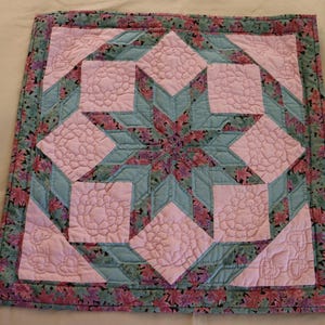 May include: A handmade quilt featuring a starburst design. The quilt has a light pink center with floral quilting, surrounded by teal and floral patterned fabric. The border is a floral print. A cozy, decorative item.
