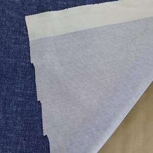 May include: A close-up of three different fabrics. The first is a textured, dark blue fabric. The second is a light blue fabric. The third is a tan fabric. The light blue fabric is folded over the dark blue fabric.