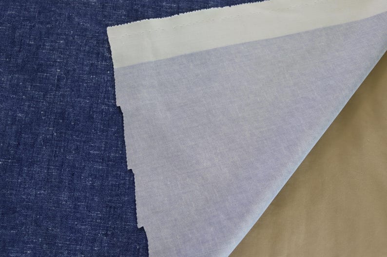 May include: A close-up of three different fabrics. The first is a textured, dark blue fabric. The second is a light blue fabric. The third is a tan fabric. The light blue fabric is folded over the dark blue fabric.