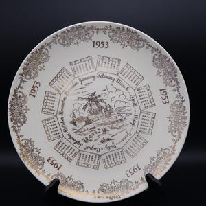 May include: A vintage decorative plate from 1953, featuring a calendar design with a windmill illustration in the center. The plate has a white background with gold-colored accents and text, including the months of the year.