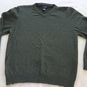 May include: A dark green V-neck sweater with long sleeves. The sweater has a ribbed hem and cuffs, and a speckled pattern throughout. The tag on the back of the neck is visible. The sweater is made of a soft, knit material.