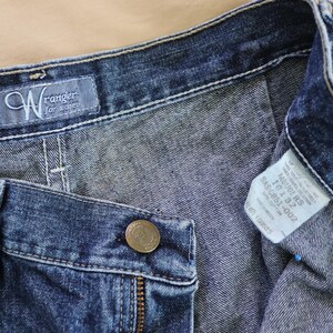 Op de afbeelding: Close-up van een blauwe denim jeans met een knoop, rits en riemlussen. De jeans heeft een "Wrangler for women" label en een maatlabel. De denim heeft een medium wassing.