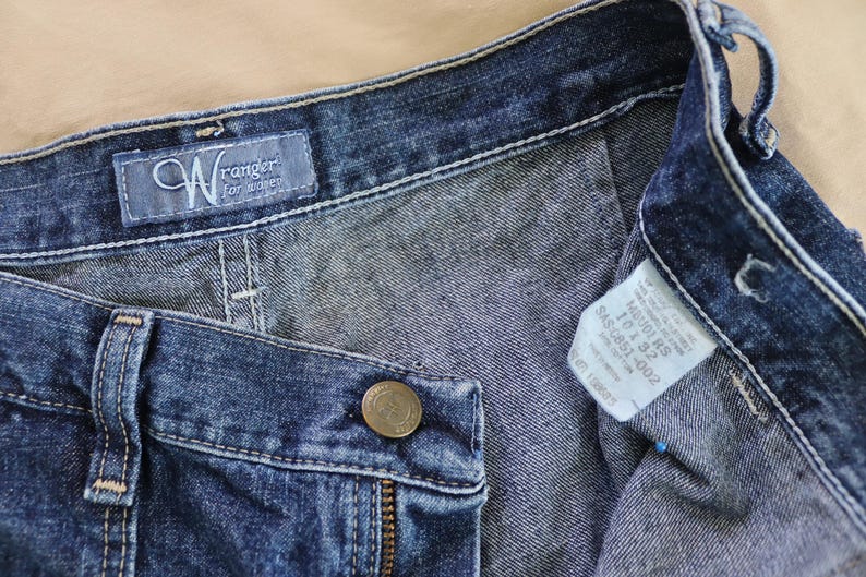 Op de afbeelding: Close-up van een blauwe denim jeans met een knoop, rits en riemlussen. De jeans heeft een "Wrangler for women" label en een maatlabel. De denim heeft een medium wassing.