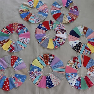 Puede incluir: Una colección de adornos circulares de tela hechos a mano. Cada adorno está compuesto por varios retazos de tela coloridos, incluyendo patrones florales, de lunares y a rayas. Los adornos están dispuestos sobre un fondo neutro, mostrando su diseño de patchwork.