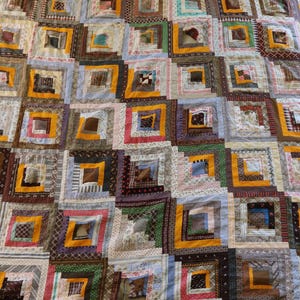 May include: A patchwork quilt with a grid pattern. Each square has a central block surrounded by strips of fabric in shades of brown, yellow, blue, and pink. The quilt is handmade, showcasing a variety of fabric patterns and colours.