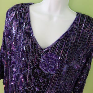 May include: A purple sequined top with a V-neck and short sleeves. The top has a floral design with a rose and leaf pattern. The fabric is embellished with sequins and beads, creating a shimmering effect. The garment is displayed on a white mannequin.