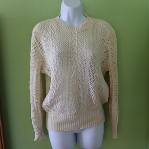 May include: Cream-colored open-knit sweater with a round neckline and long sleeves. The sweater features a textured diamond pattern on the front and sleeves, and a ribbed hem and cuffs. The sweater is displayed on a white mannequin.