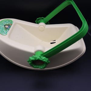 May include: A white plastic toy basket with a green handle and accents. The basket has a curved shape and a logo on the side that says "Patch Kids". The handle is attached with white and green plastic pieces.