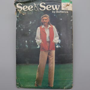 May include: Vintage See & Sew Butterick pattern envelope featuring a woman in a red vest, white shirt, and tan pants. The envelope has the text "See & Sew" and "Butterick" printed on it. The background is a blurred green and brown.