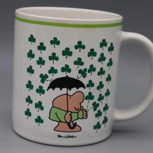May include: White ceramic mug with a green rim and handle. The mug features a cartoon illustration of a person holding a black umbrella under a shower of green shamrocks. The artist's signature is at the bottom.