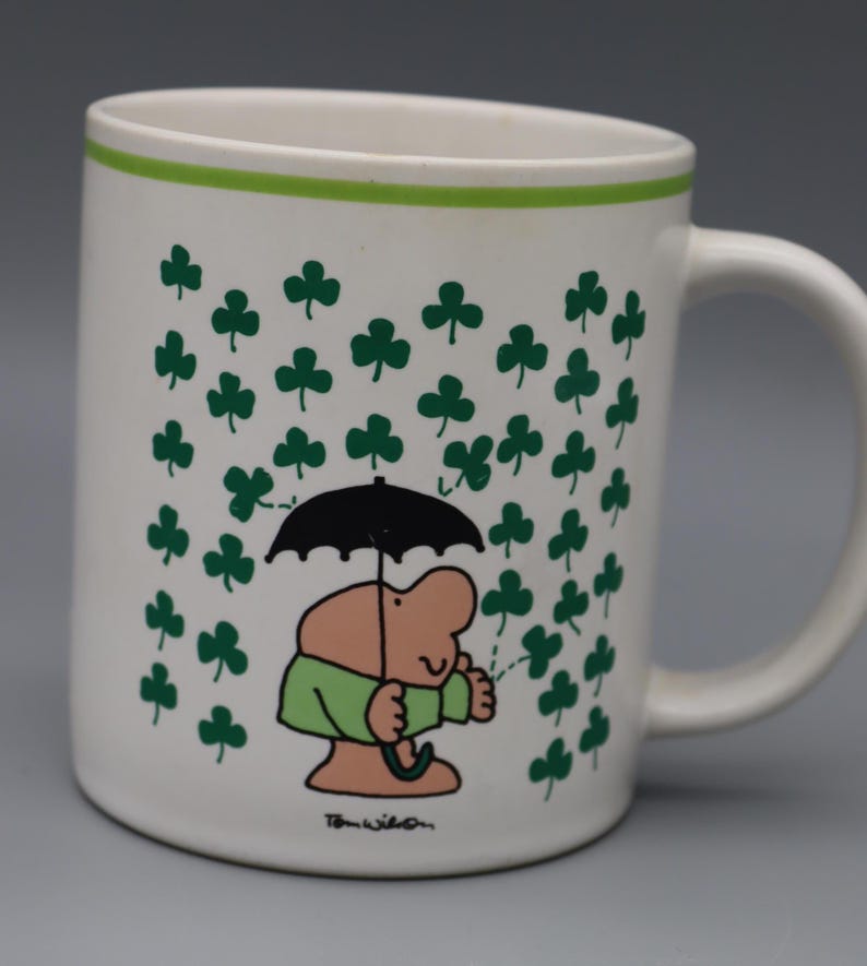 May include: White ceramic mug with a green rim and handle. The mug features a cartoon illustration of a person holding a black umbrella under a shower of green shamrocks. The artist's signature is at the bottom.