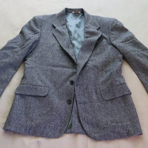 Puede incluir: Una blazer de tweed gris con cierre de dos botones, solapas con muescas y dos bolsillos delanteros. El forro interior es de color azul claro. Las mangas tienen tres botones en los puños. Esta chaqueta clásica es adecuada para varias ocasiones.