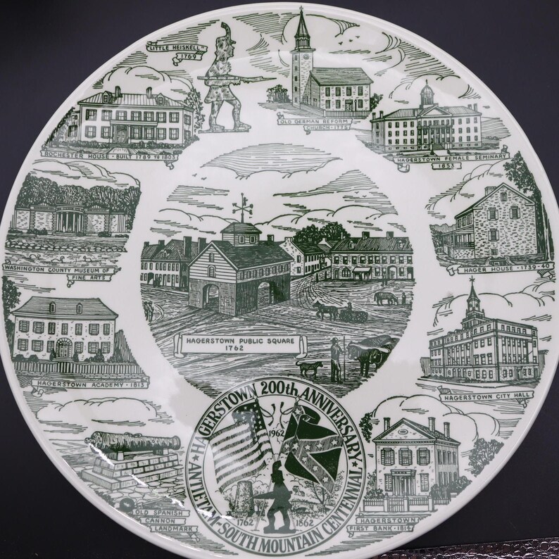K&ouml;nnte beinhalten: Ein wei&szlig;er Keramikteller mit einer gr&uuml;nen Illustration von Wahrzeichen von Hagerstown, Maryland. Der Teller zeigt Geb&auml;ude, einen Soldaten, Flaggen und den Text "Hagerstown 200th Anniversary" und "Antietam South Mountain Centennial".