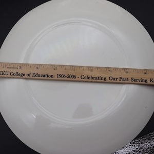K&ouml;nnte beinhalten: Ein wei&szlig;er Keramikteller mit leicht erh&ouml;htem Rand. Ein Holzlineal, in Zoll markiert, liegt auf dem Teller. Auf dem Lineal steht "EKU College of Education: 1906-2006 - Celebrating Our Past: Serving Kentucky".