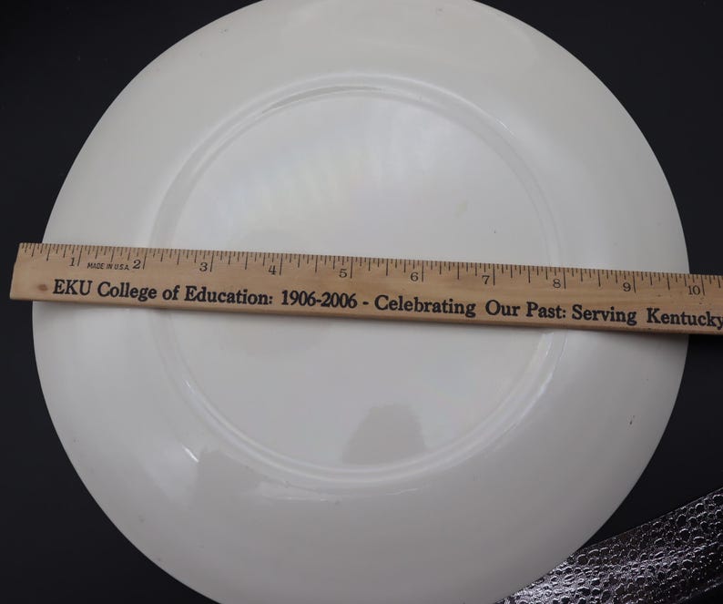 K&ouml;nnte beinhalten: Ein wei&szlig;er Keramikteller mit leicht erh&ouml;htem Rand. Ein Holzlineal, in Zoll markiert, liegt auf dem Teller. Auf dem Lineal steht "EKU College of Education: 1906-2006 - Celebrating Our Past: Serving Kentucky".