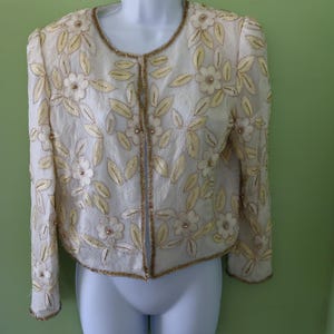 May include: A white jacket with gold embroidery of flowers and leaves. The jacket has a round neckline and long sleeves.