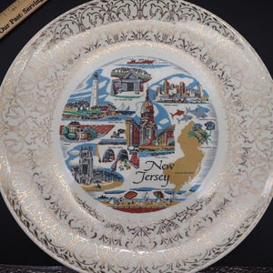 K&ouml;nnte beinhalten: Ein dekorativer Vintage-Teller mit einer Karte von New Jersey mit ikonischen Wahrzeichen und Illustrationen. Der Teller hat einen wei&szlig;en Hintergrund mit goldenen Filigranarbeiten am Rand und einem goldenen Rand. Die Mitte des Tellers zeigt die Landkarte des Staates.