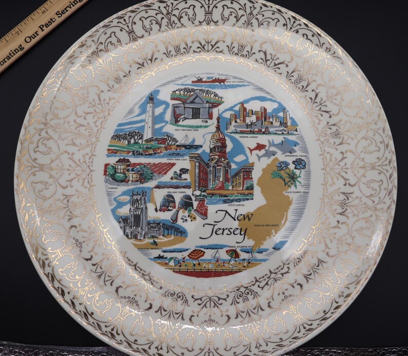 K&ouml;nnte beinhalten: Ein dekorativer Vintage-Teller mit einer Karte von New Jersey mit ikonischen Wahrzeichen und Illustrationen. Der Teller hat einen wei&szlig;en Hintergrund mit goldenen Filigranarbeiten am Rand und einem goldenen Rand. Die Mitte des Tellers zeigt die Landkarte des Staates.