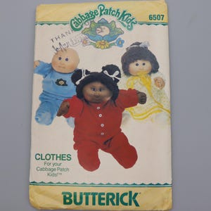 May include: A Butterick sewing pattern envelope for Cabbage Patch Kids clothes, number 6507. The front features illustrations of three dolls wearing a blue outfit, a red jumpsuit, and a yellow dress. The text includes Cabbage Patch Kids and Butterick.