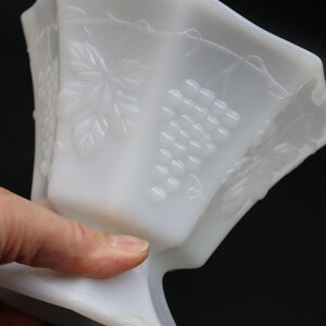 May include: A white, vintage-style glass bowl with a pedestal base. The bowl features embossed grape and leaf designs on its sides. The bowl is held by a hand, showcasing its shape and details against a dark background.